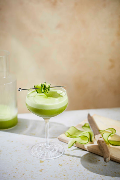 A bright green Verdant Margarita in a coupe with a cucumber garnish