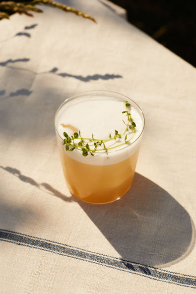 A Whiskey Sour in a tumbler with foam and thyme, on sunlit linen