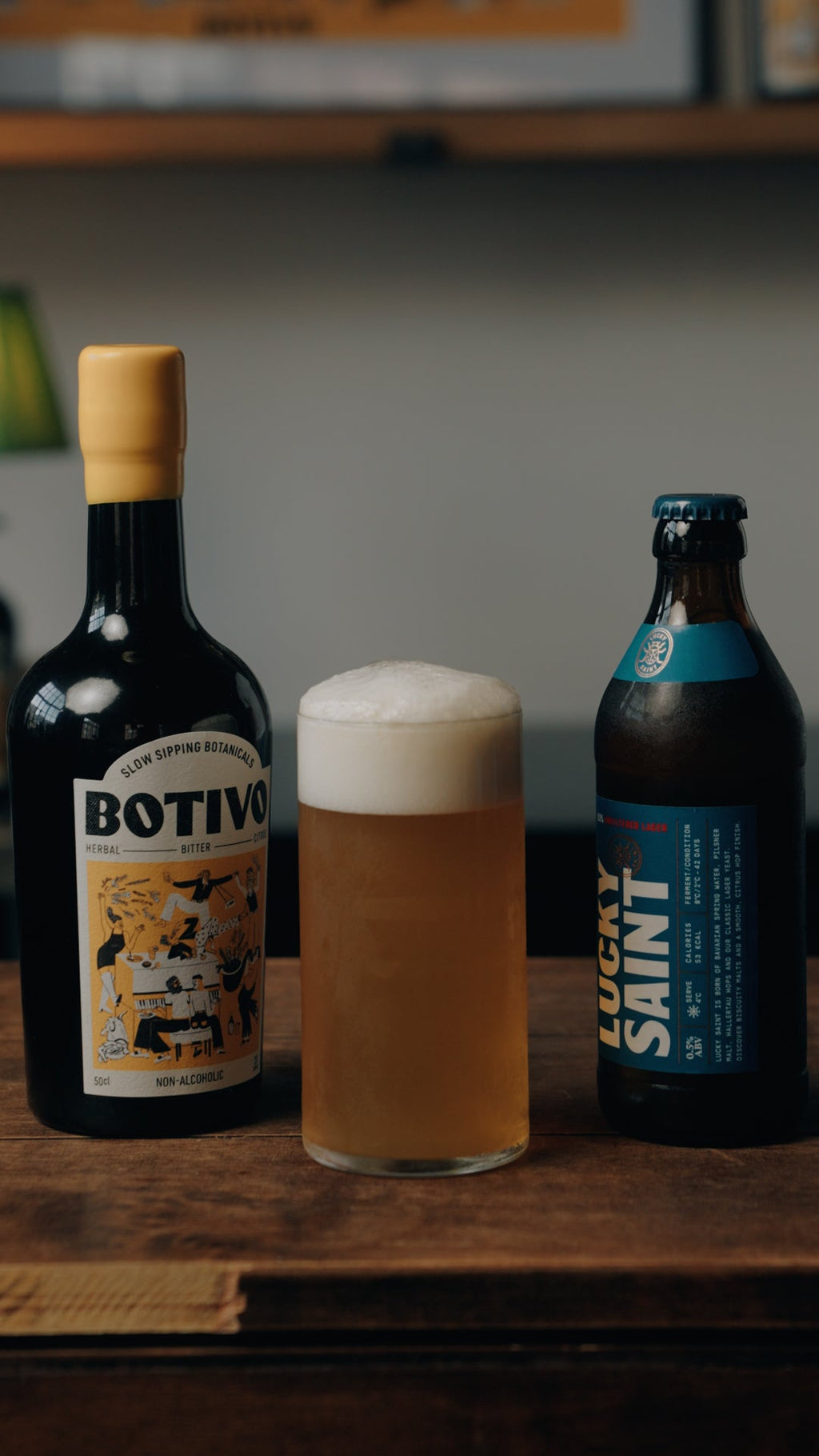 A bottle of Botivo and a bottle of Lucky Saint beside a tall shandy with a thick head