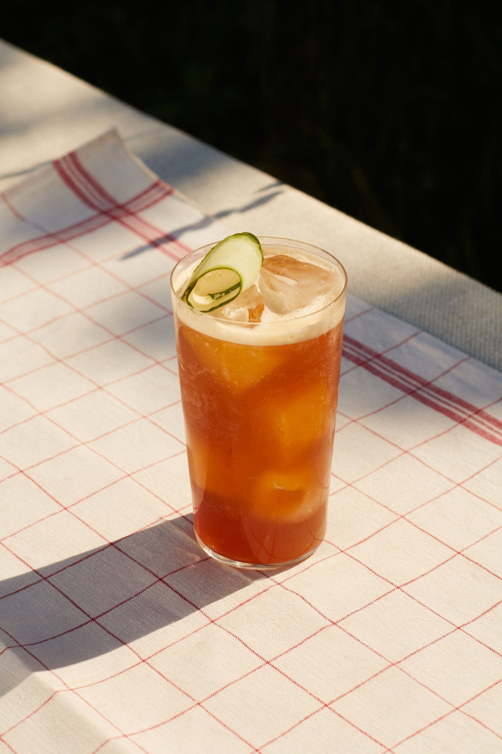 A tall glass of deep red Bitter Red topped with cucumber slices on a checked tea towel
