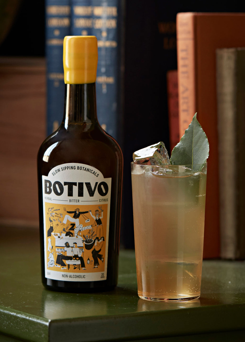 A bottle of Botivo beside a Whiskey Highball with a bay leaf, books in the background