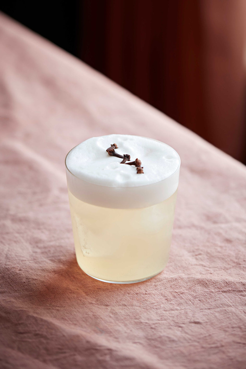 A Clove Sour in a tumbler with thick foam and cloves scattered on top, on a pink cloth