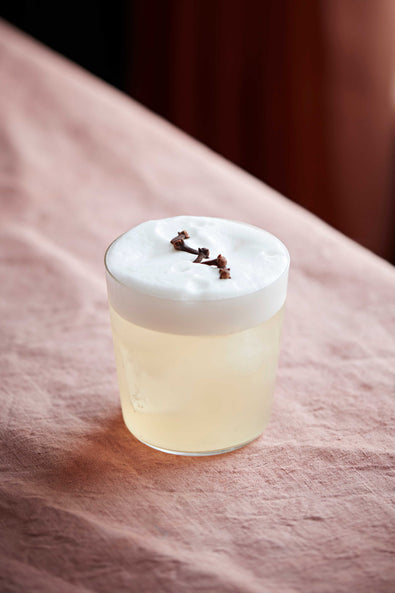  A Clove Sour in a tumbler with thick foam and cloves scattered on top, on a pink cloth 