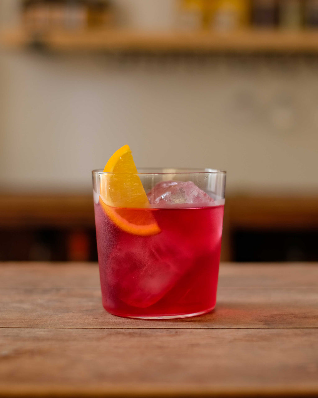A deep red Botivo Nogroni in a tumbler over ice with an orange wedge on a wooden bar