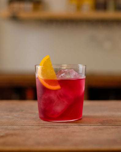  A deep red Botivo Nogroni in a tumbler over ice with an orange wedge on a wooden bar 