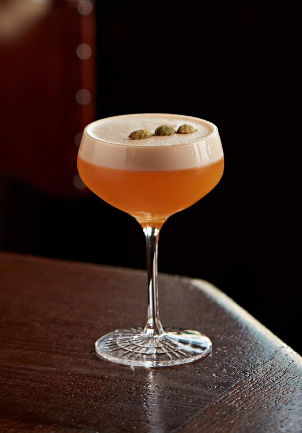 A Clementine Sour in a coupe topped with cardamom pods on a dark bar
