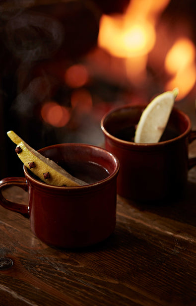 Two enamel mugs of Hot Botty with clove-studded lemon wedges, by a fire