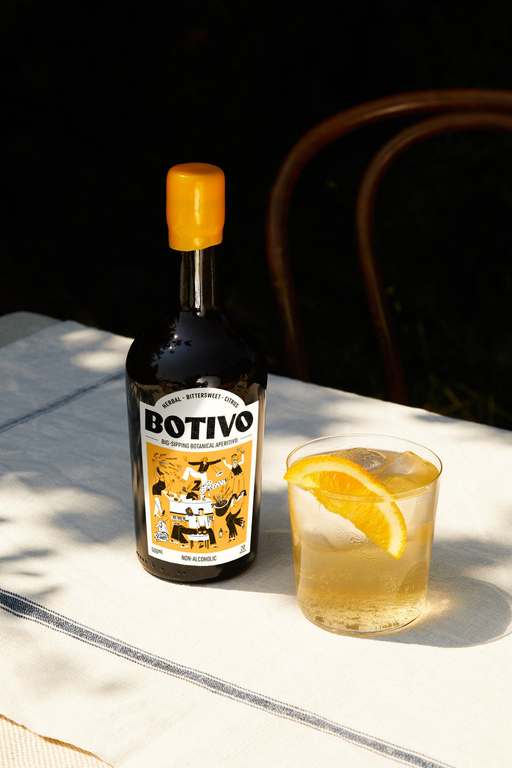 A bottle of Botivo beside a glass of Botivo and soda with an orange wedge, on a sunlit table