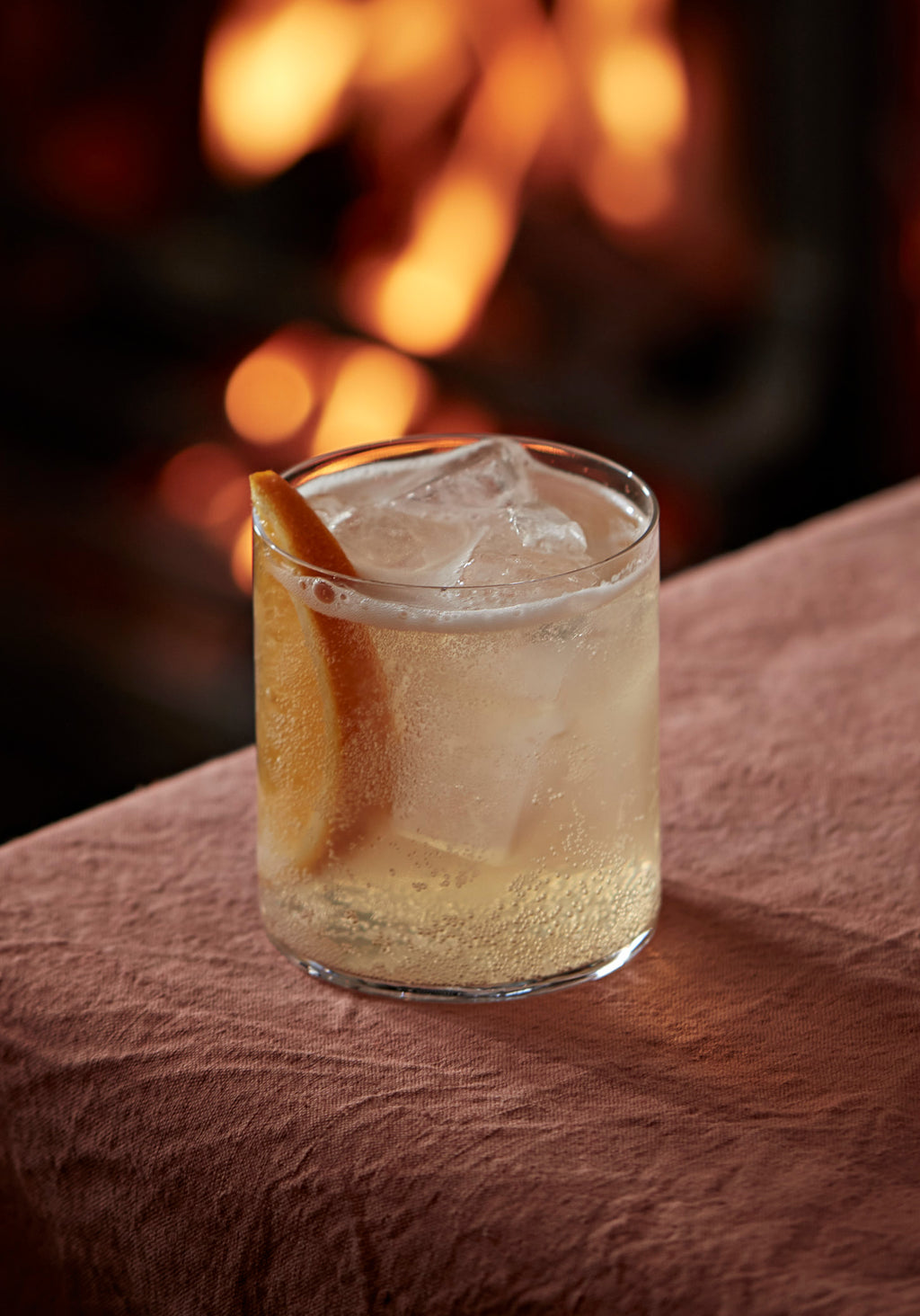 A tumbler of Botivo and tonic over ice with an orange wedge, firelight behind