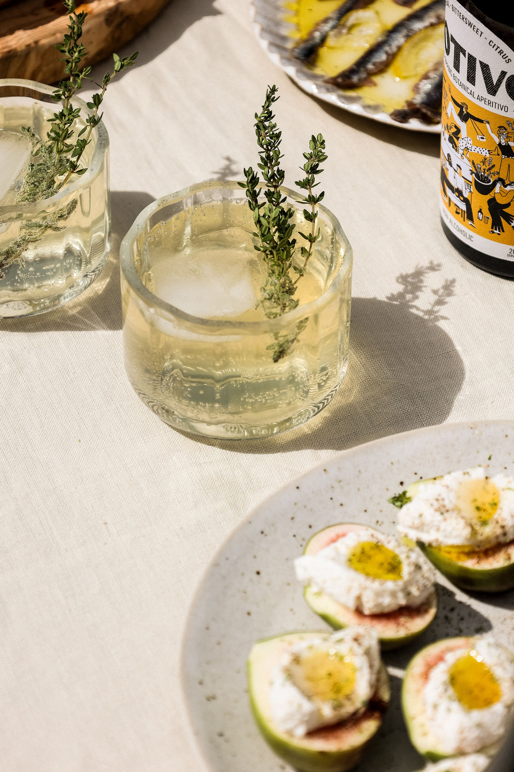 A tumbler of EVOO Botivo with a thyme sprig, beside figs and a plate of olive oil