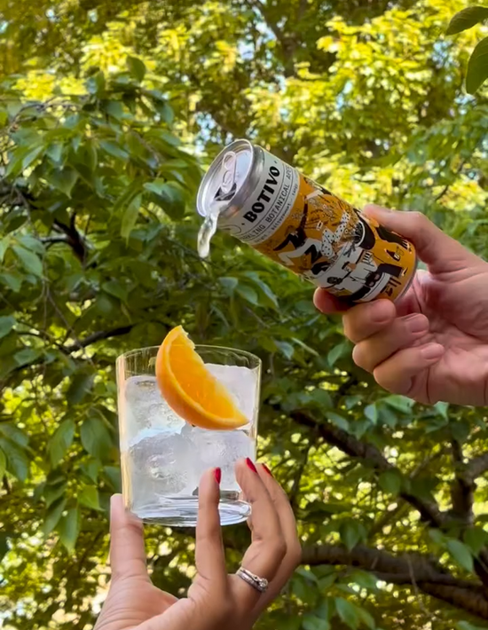  Can of Botivo and soda being poured into a glass with ice and an orange slice under green trees 