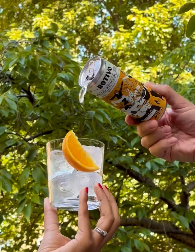  Can of Botivo and soda being poured into a glass with ice and an orange slice under green trees 